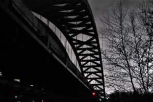 Contre-plongée nocturne sur l’arche métallique d’un pont routier se découpant en silhouette sur un ciel gris, arbres nus à droite et petits feux de signalisation rouges et verts en bas