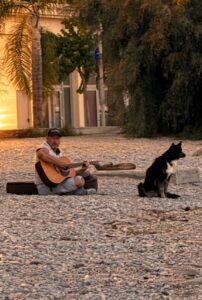 Homme jouant de la guitare assis sur une plage de galets, chien noir à ses côtés, dans une lumière dorée de fin de journée