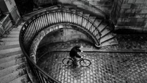 Vue plongeante en noir et blanc sur un escalier de pierre en courbe à la rampe de fer forgé ; en bas, un cycliste traverse des pavés mouillés.