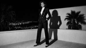 Femme assurée en tailleur-pantalon croisé et escarpins sur un toit-terrasse de la Riviera la nuit, flash direct et grande ombre portée sur un mur blanc, noir et blanc contrasté.