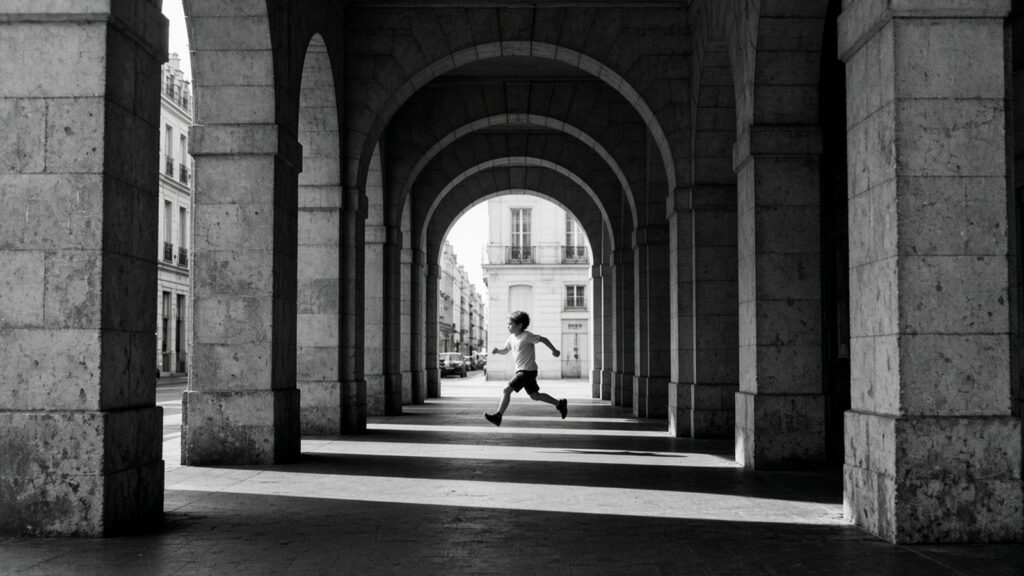 Un enfant court, les deux pieds en l’air, sous une enfilade d’arcades de pierre baignées de soleil, en noir et blanc.